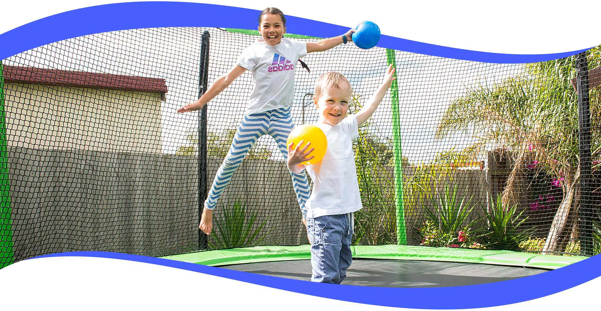 A young girl and a little boy smiling, holding rubber balls and bouncing on a large outdoor trampoline with a safety net enclosure